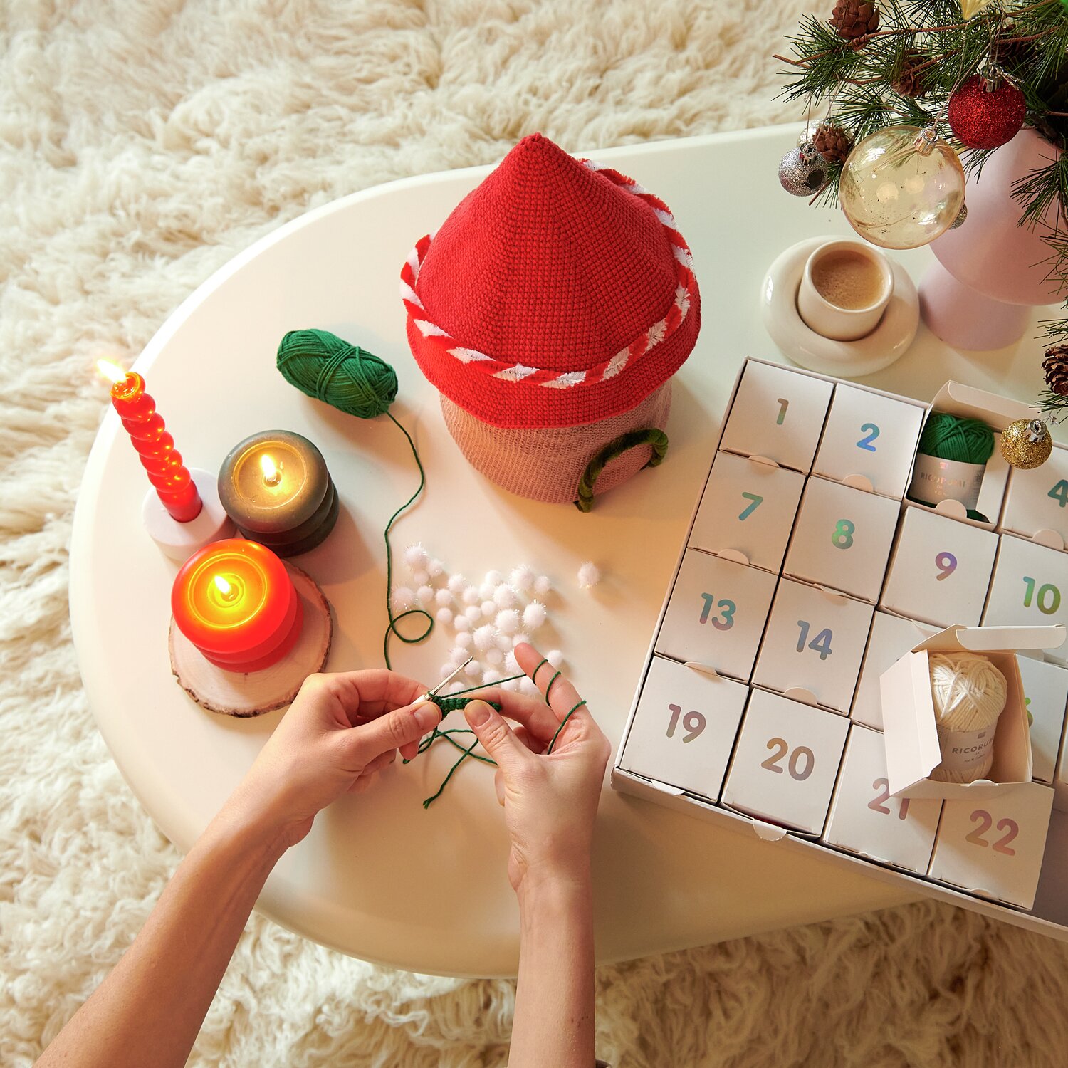 Ricorumi Adventskalender: Hände basteln mit grünem Garn neben Adventskalender und roten Kerzen auf weißem Tisch.