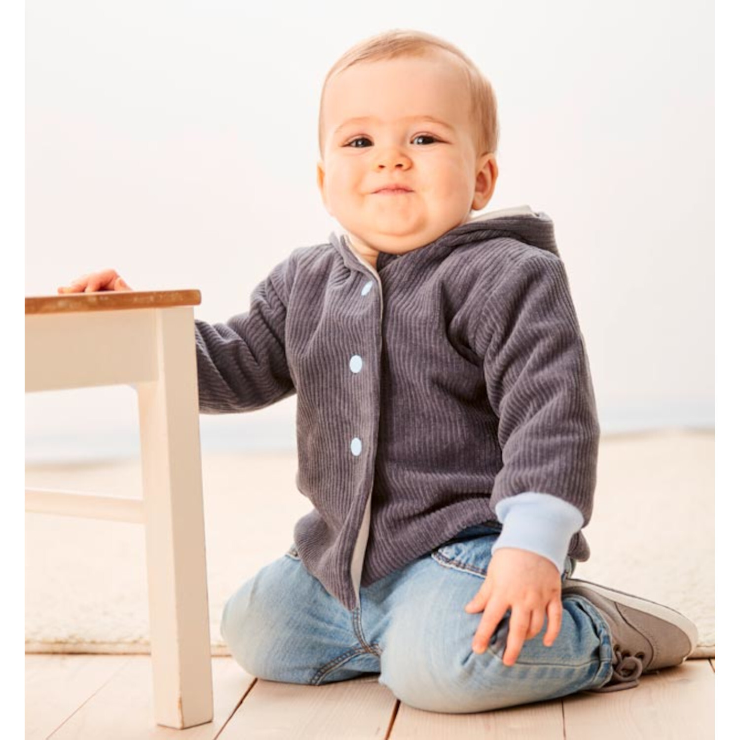 Nähen mit Jersey für Babys: Ein lächelndes Baby in blauer Kapuzenjacke und Jeans sitzt auf hellem Holzboden.