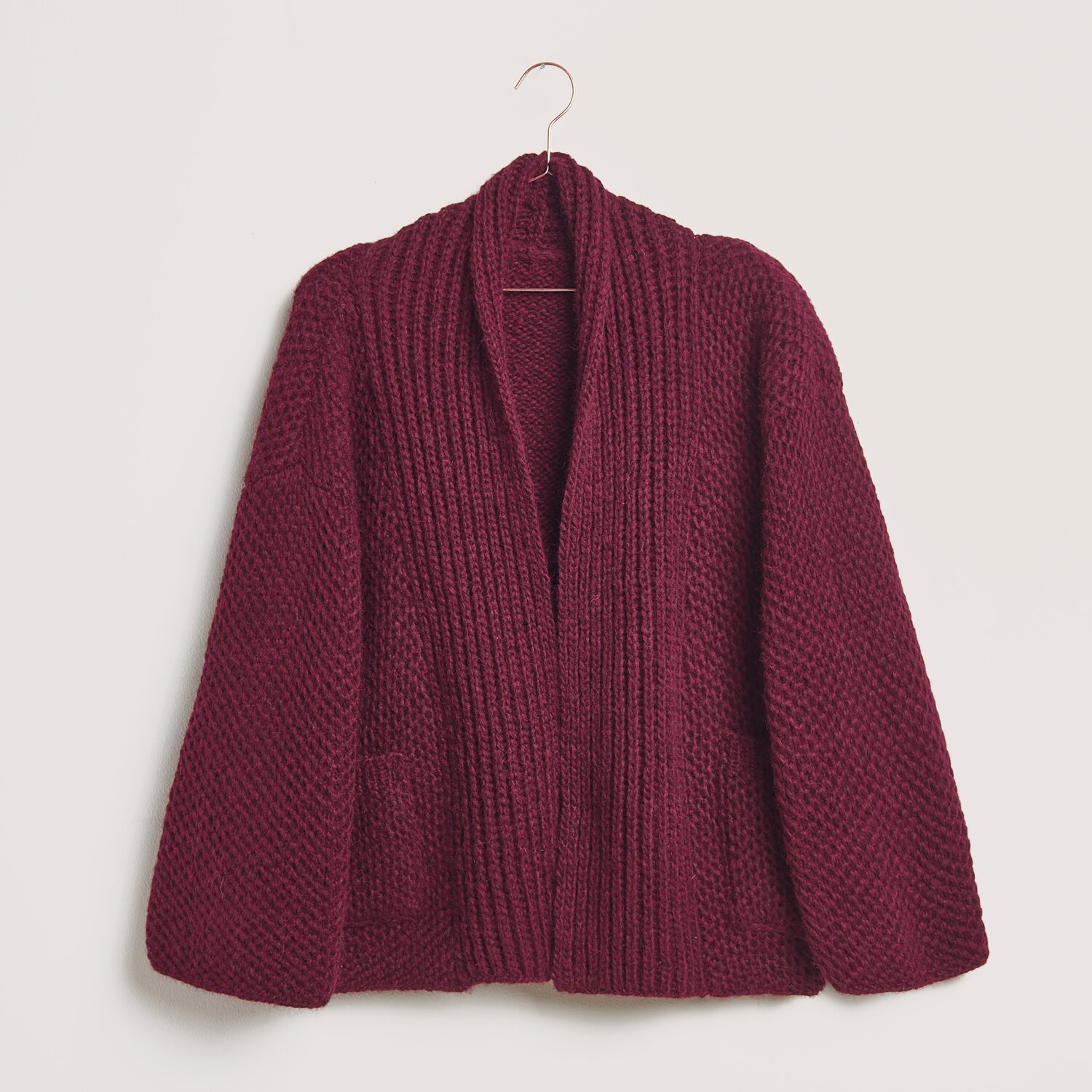 Made by Me No. 21 Herbst-Winter Sprachauswahl deutsch von Rico Design Stickset Jacke Modell 29 aus Made by Me No. 21: Bordeauxfarbene Strickjacke aus Alpaka-Merinowolle, präsentiert auf einem Kleiderbügel.
