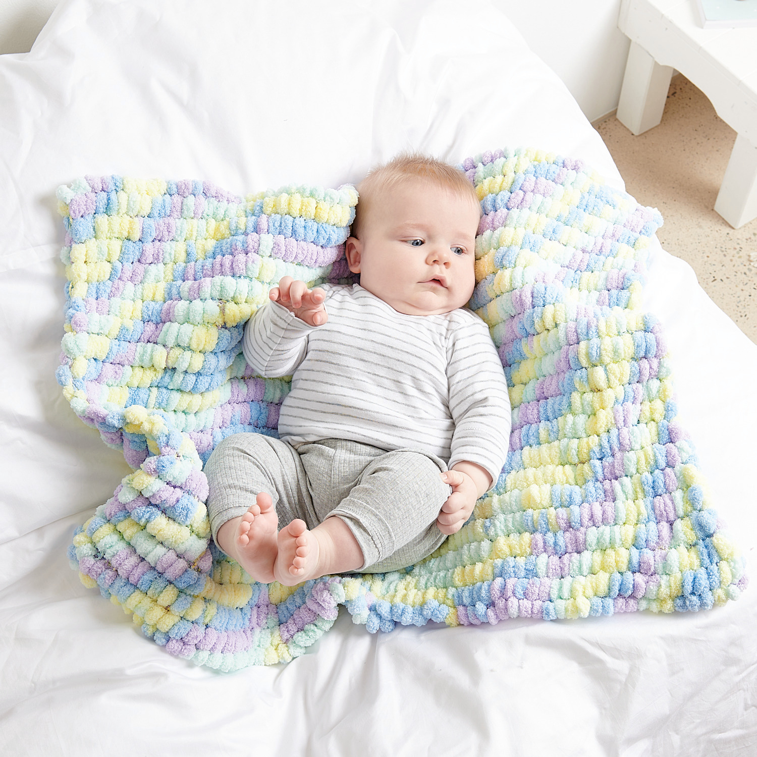 Creative Pompon Print 200g Farbe Blau Bunt von Rico Design Creative Pompon Print: Ein Baby liegt auf einer weichen, bunten Decke von Rico Design in Blau Bunt.