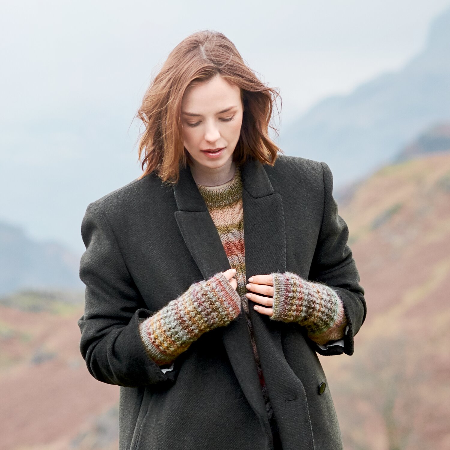 Strickset Stulpen Modell 38 aus Lovewool No. 23, warme gestrickte Handstulpen im Naturmelange-Look, getragen von Frau im Mantel vor nebliger Berglandschaft