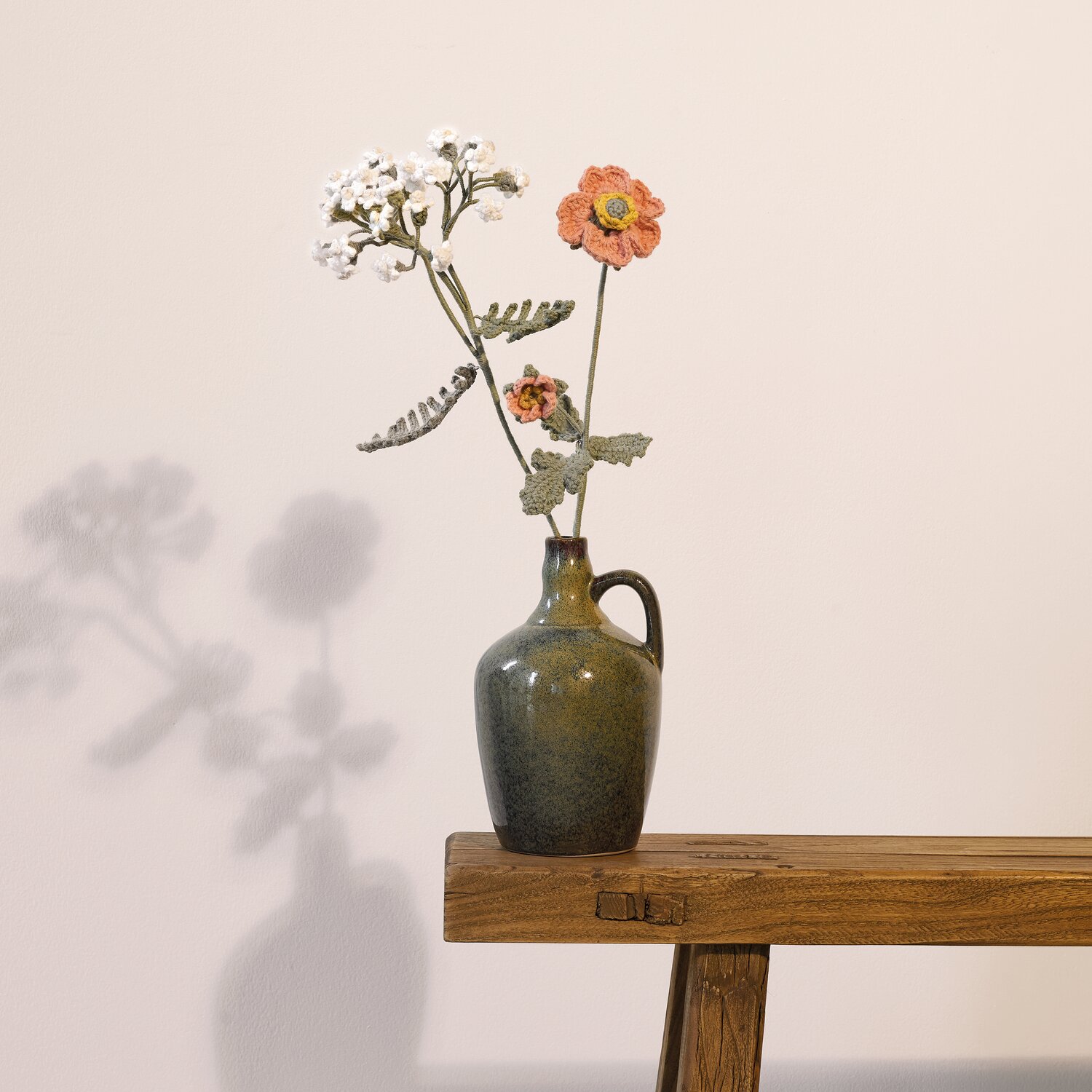 Ricorumi In the Meadow arrangiert gehäkelte Blumen in gedeckten Farben in einer grünen Keramikvase auf rustikalem Holztisch vor heller Wand.