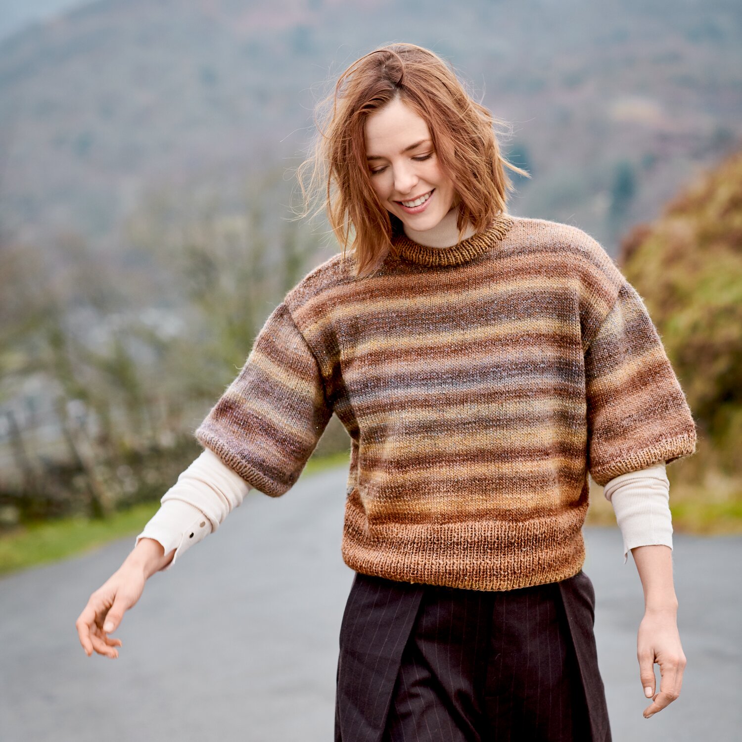 Strickset Shirt Modell 35 aus Lovewool No. 23, kurzärmliges gestreiftes Oberteil in warmen Brauntönen, getragen von Frau beim Spaziergang in herbstlicher Landschaft