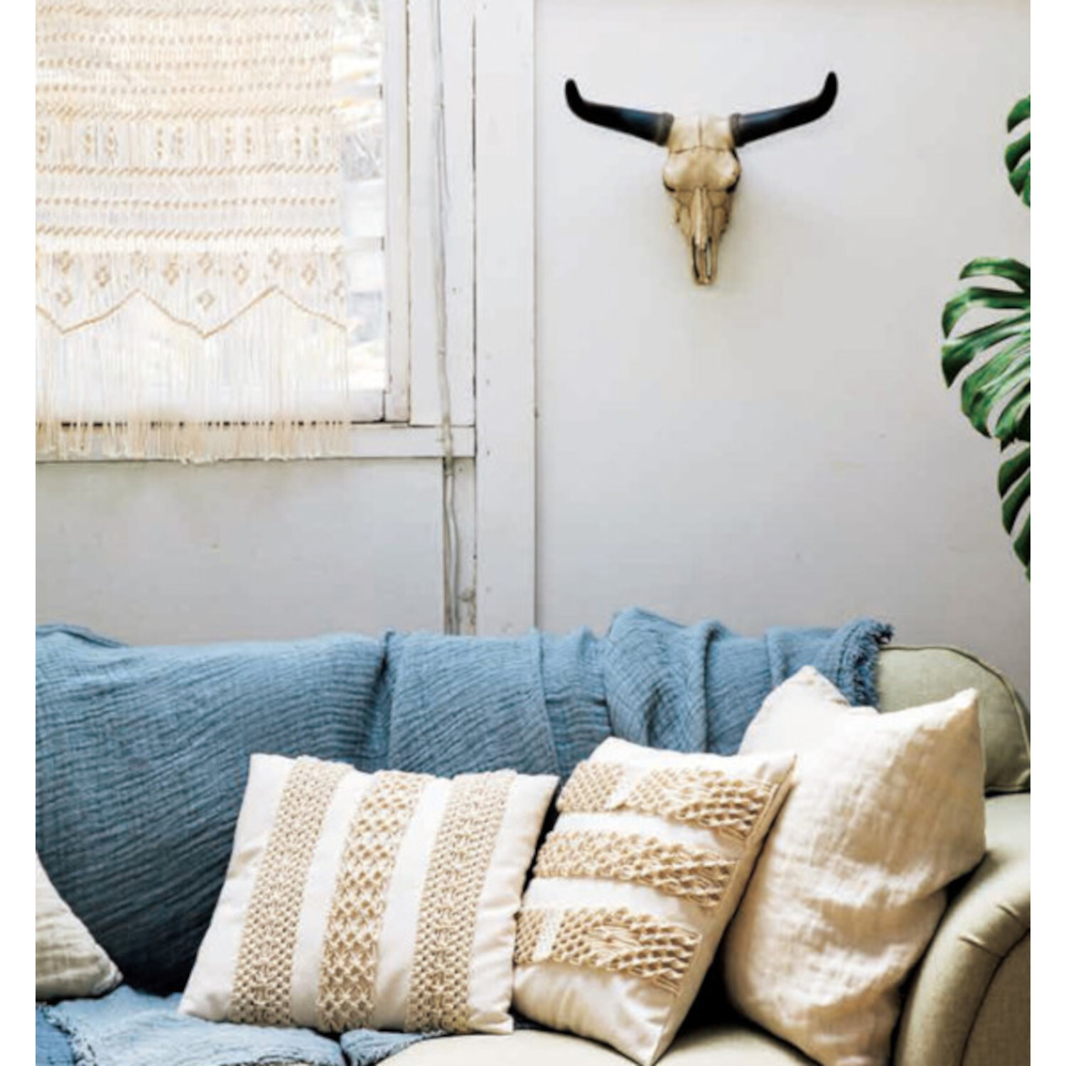 New Makramee - 25 Knüpfprojekte: Gemütliches Wohnzimmer mit blauen und beigen Kissen auf einem Sofa, dekoriert mit Makramee und Stierschädel an der Wand.