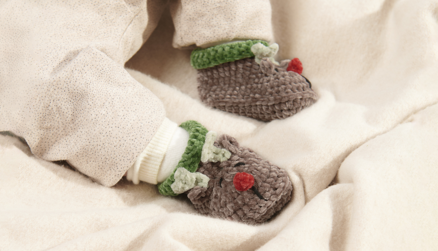 Häkelanleitung Baby Booties Rentier: Niedliche braune Babyschuhe mit Rentiermotiv und grünen Details auf beigem Stoff.