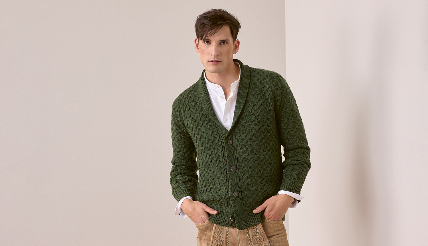 Strickanleitung Trachtenjacke aus Essentials Merino Plus dk Trachtenjacke aus Essentials Merino Plus dk, grün, mit Knopfleiste, getragen von einem Mann vor neutralem Hintergrund.