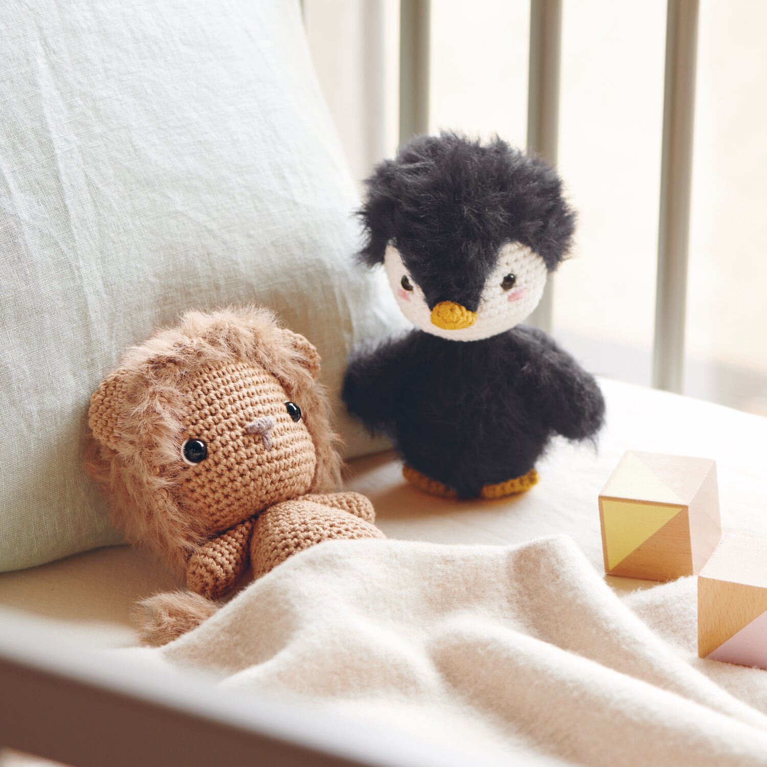 Creative Ricorumi dk 25g Farbe Creme von Rico Design Ricorumi Furry Animals: Kuscheltier-Löwe in Karamell und Pinguin in Schwarz auf Bett mit Holzklötzen.