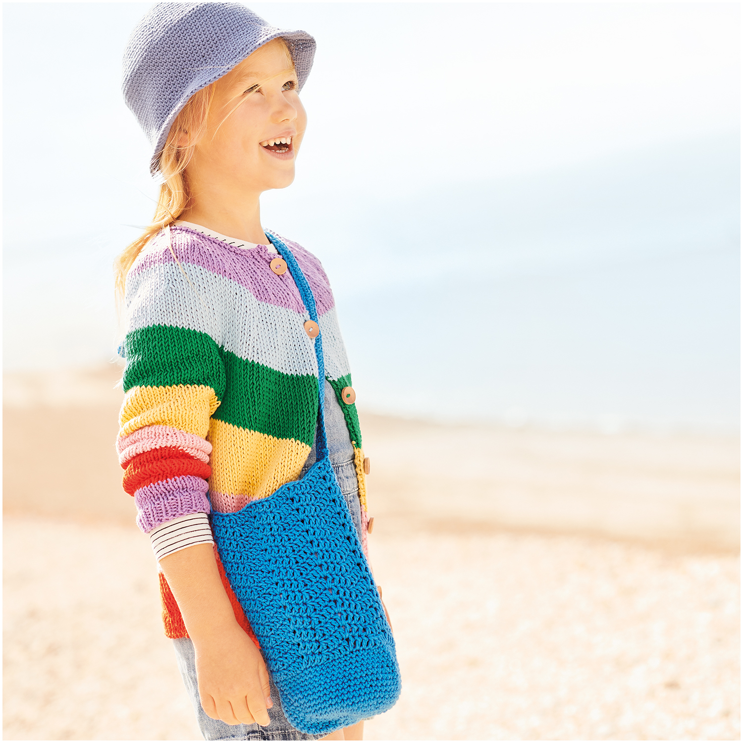 Essentials Organic Cotton aran: Kind am Strand mit bunter Strickjacke und himmelblauer Tasche.