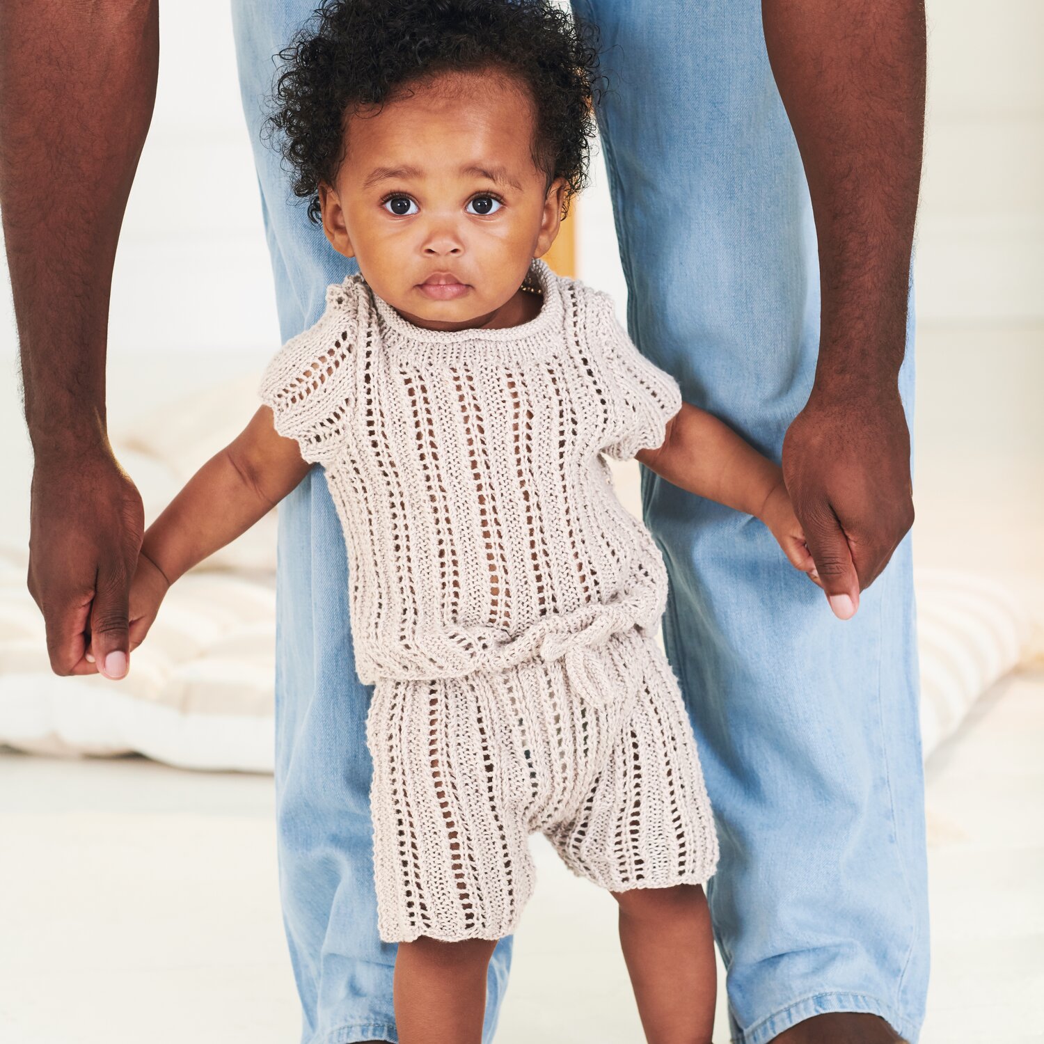 Strickset Shirt Modell 17 aus Baby 043: Baby in cremefarbenem Strickoutfit, gehalten von einem Erwachsenen in Jeans.