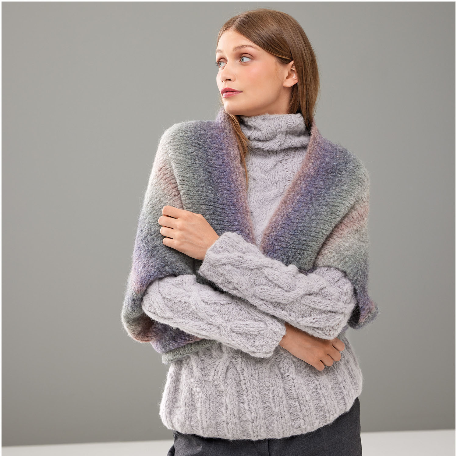 Strickset Poncho Modell 29 aus Die Neue Masche Nr. 8 von idee. Creativmarkt Strickset Poncho Modell 29 aus Die Neue Masche Nr. 8: Frau trägt grauen Rollkragenpullover und bunten Poncho vor grauem Hintergrund.