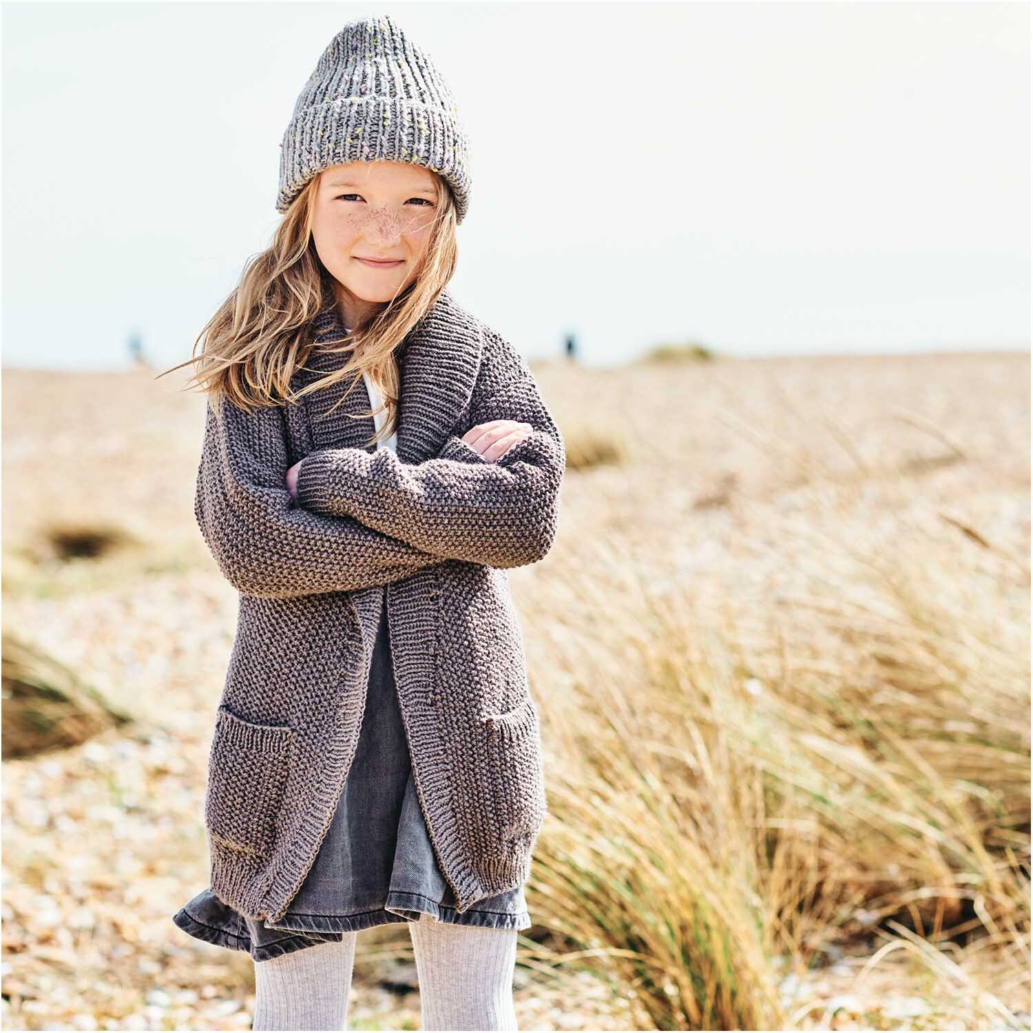 Essentials Super aran: Mädchen in gestricktem Outfit am Strand, trägt grauen Cardigan und Mütze von Rico Design.