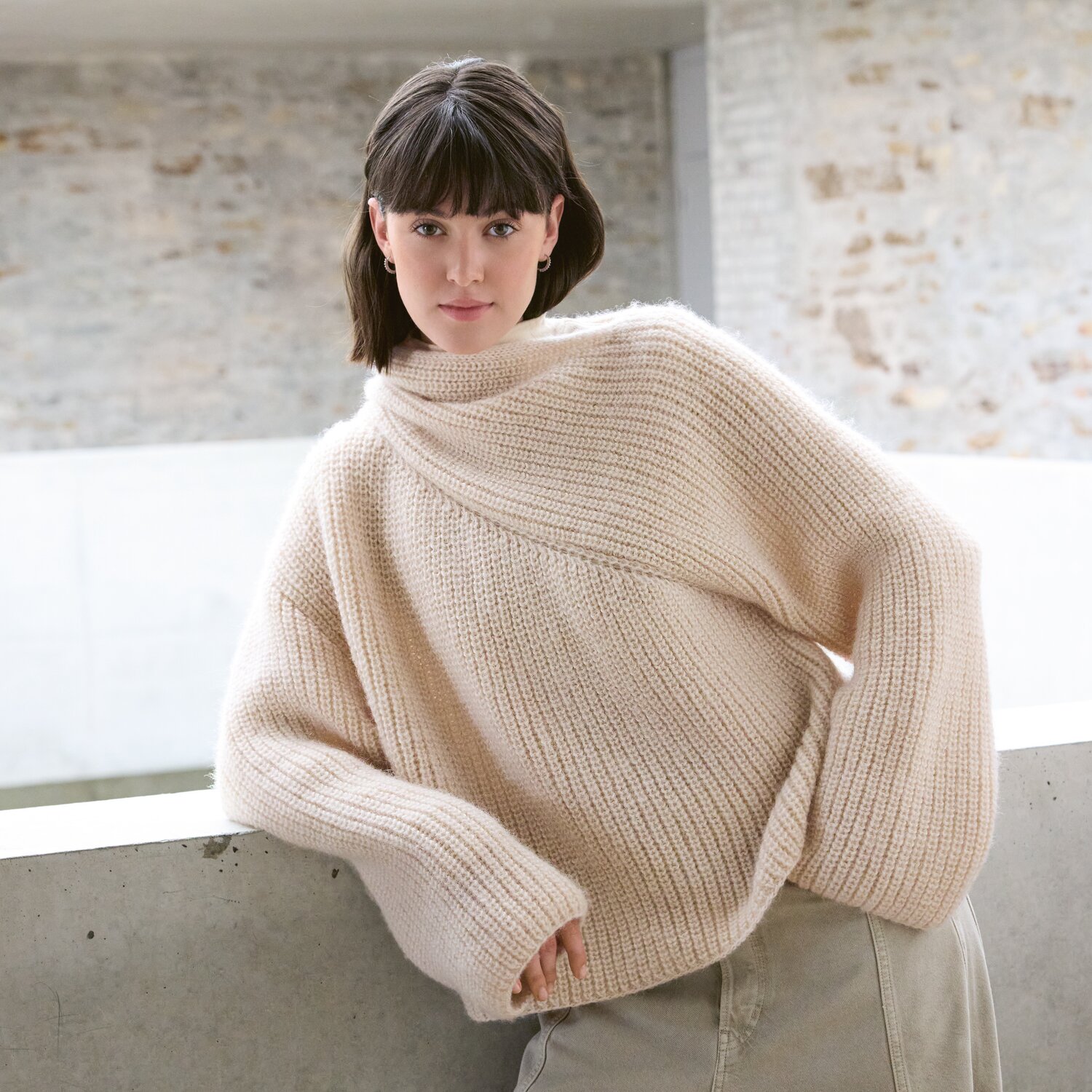 Strickset Pullover Modell 01 aus Luxury Knits zeigt eine Frau in einem cremefarbenen Pullover vor einer Steinwand.