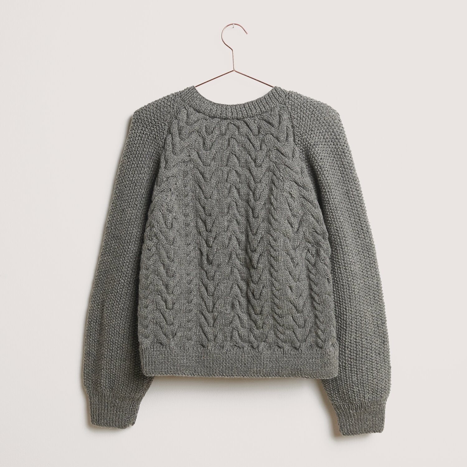 Essentials Soft Merino aran 50g 100m Farbe Mittelgrau von Rico Design Essentials Soft Merino aran Pullover in Mittelgrau, gestrickt aus Rico Design Wolle, hängt an einem Kleiderbügel vor neutralem Hintergrund.