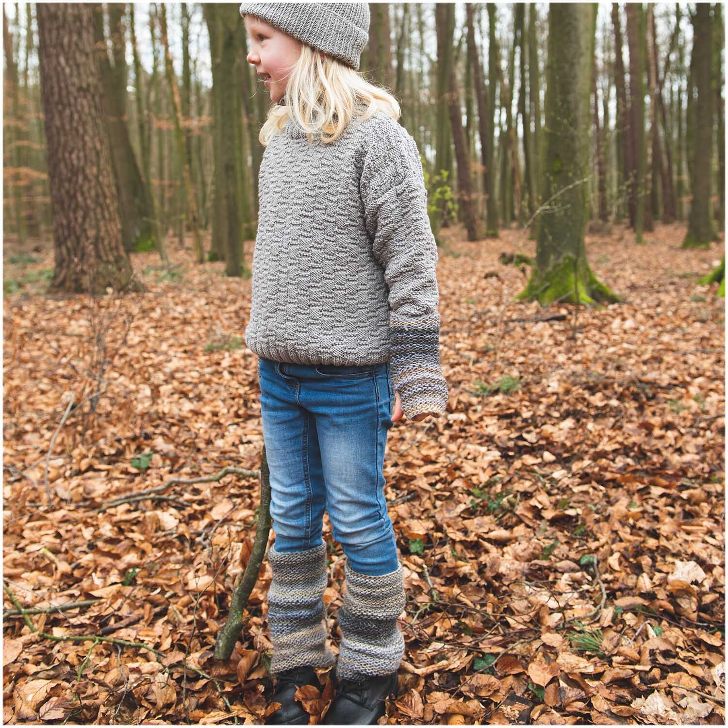 Essentials Merino dk: Kind im gestrickten Outfit im Wald, umgeben von Herbstlaub, trägt grauen Pullover und Mütze.