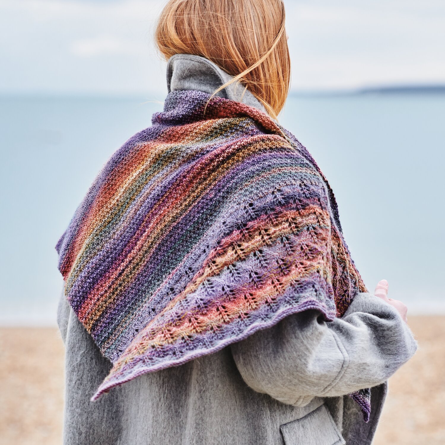 Strickset Tuch Modell 08 aus Wonderball Special von Rico Design Strickset Tuch Modell 08 aus Wonderball Special: Frau am Strand mit buntem, gestricktem Tuch in Lila- und Brauntönen über grauem Mantel.