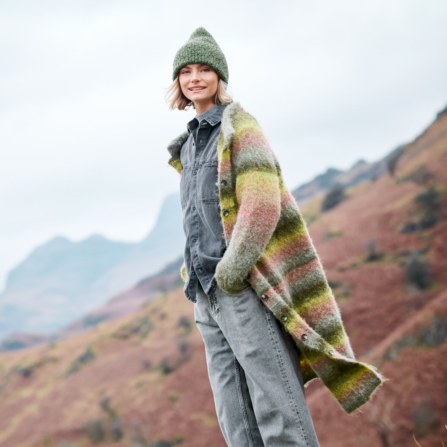 Strickset Jacke Modell 21 aus Lovewool No. 23, lang geschnittener Cardigan in mehrfarbigem Melange-Garn, getragen von Frau mit passender Mütze in herbstlicher Berglandschaft