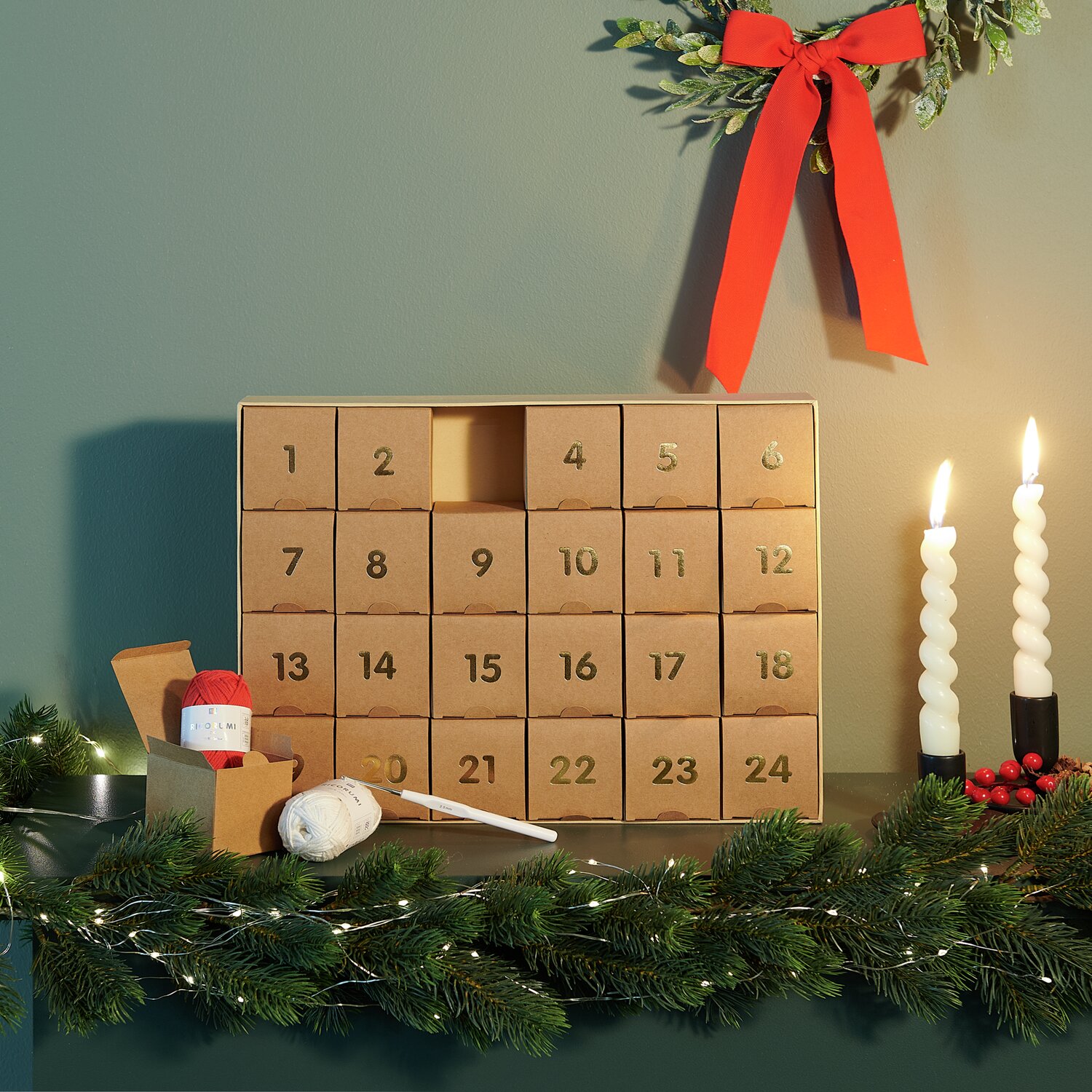 Ricorumi Adventskalender mit 24 nummerierten Fächern, dekoriert mit Kerzen und Tannenzweigen.