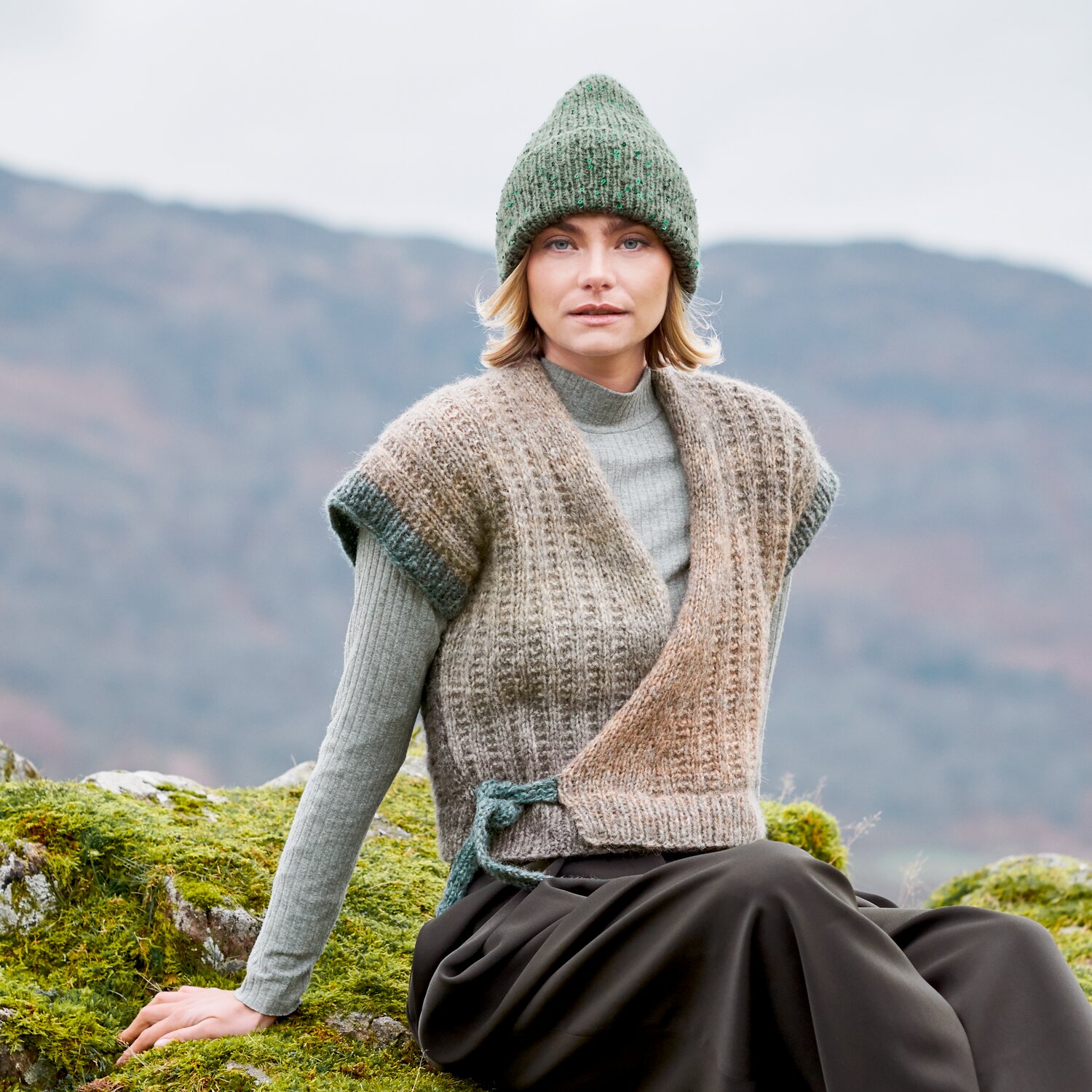 Strickset Weste Modell 15 aus Lovewool No. 23, lässig gemusterte Wickelweste, von Frau mit Mütze im Naturpanorama getragen, ideal für gemütliche Outdoor-Looks