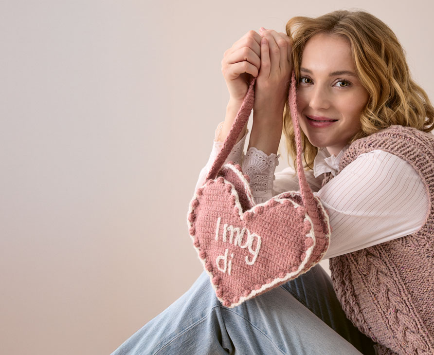 Gehäkelte Tasche in Herzform mit der Aufschrift "I mog di" wird von einem Model gehalten.