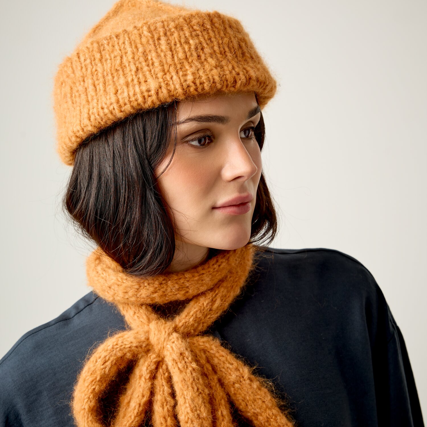 Strickset Mütze 13 aus Luxury Knits Little Pieces, grob gestrickte Mütze und Schal in warmem Senfton, getragen von Frau in dunkelblauer Bluse