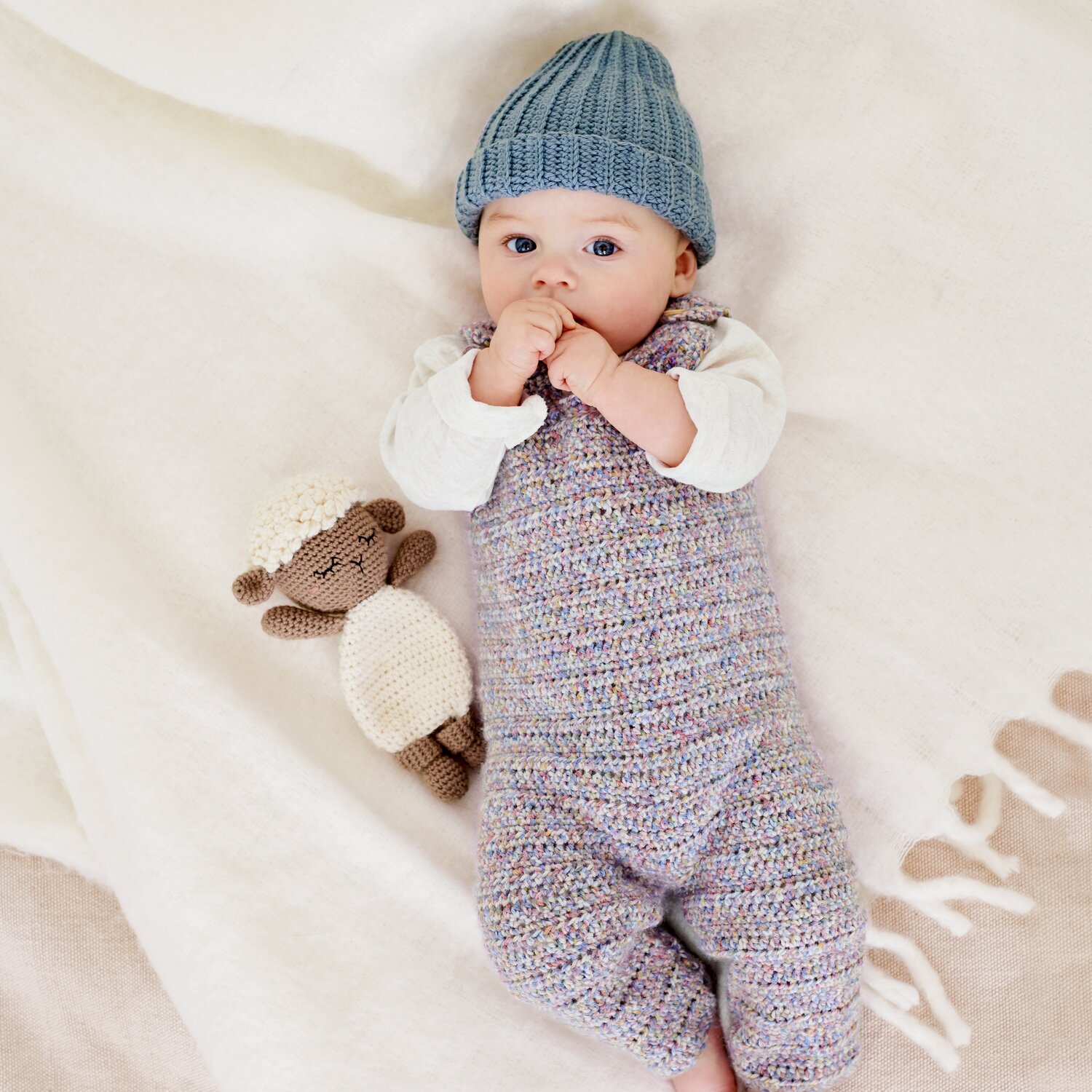Häkelset Strampler Modell 18 aus Baby 044, gemütlicher Einteiler aus mehrfarbigem Garn, getragen von liegendem Baby auf Decke neben gehäkeltem Schafsspielzeug