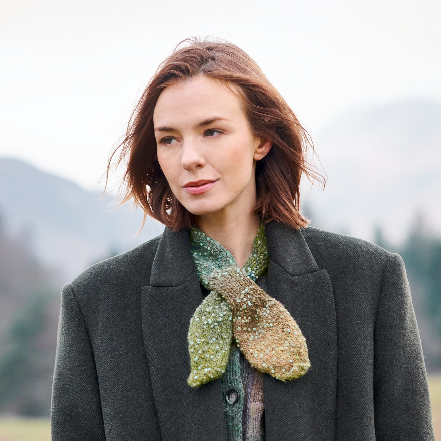 Strickset Minituch Modell 22 aus Lovewool No. 23, gestricktes Minituch im Naturlook, locker um den Hals einer Frau im dunklen Mantel vor Landschaft gebunden