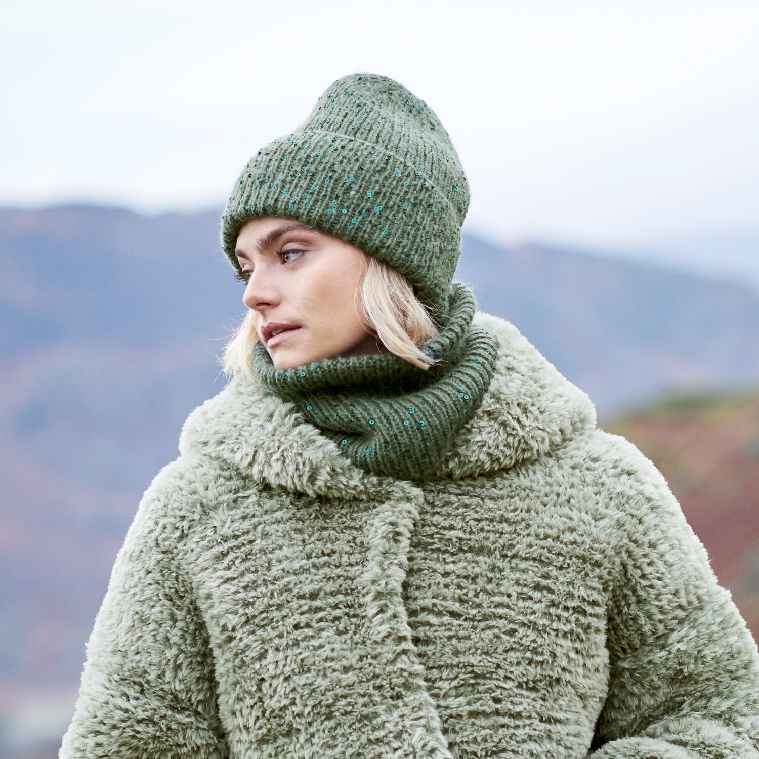 Strickset Loop Modell 20 aus Lovewool No. 23, gestrickter Loop mit passender Mütze, getragen von Frau im Freien mit winterlicher Berglandschaft im Hintergrund