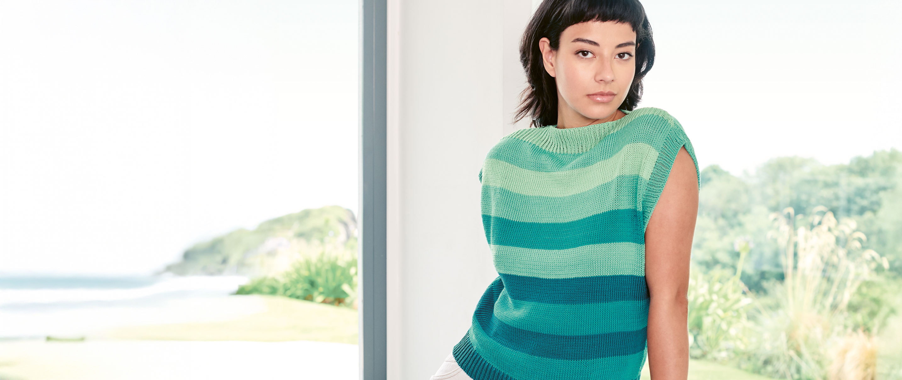 Stricktop in Aquatönen mit Blockstreifen wird von einem Model getragen vor einer Meeres-Kulisse.