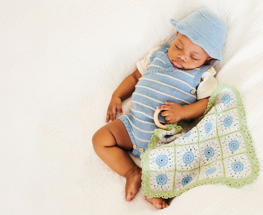 Baby liegt in Strickstrampler mit Häkeldecke auf Bett.