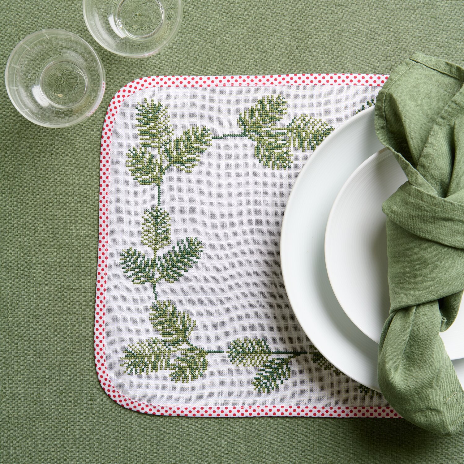 Stickpackung Platzsets mit Tannenzweigen, 2 Stück, von Rico Design. Auf grünem Tisch mit Geschirr.