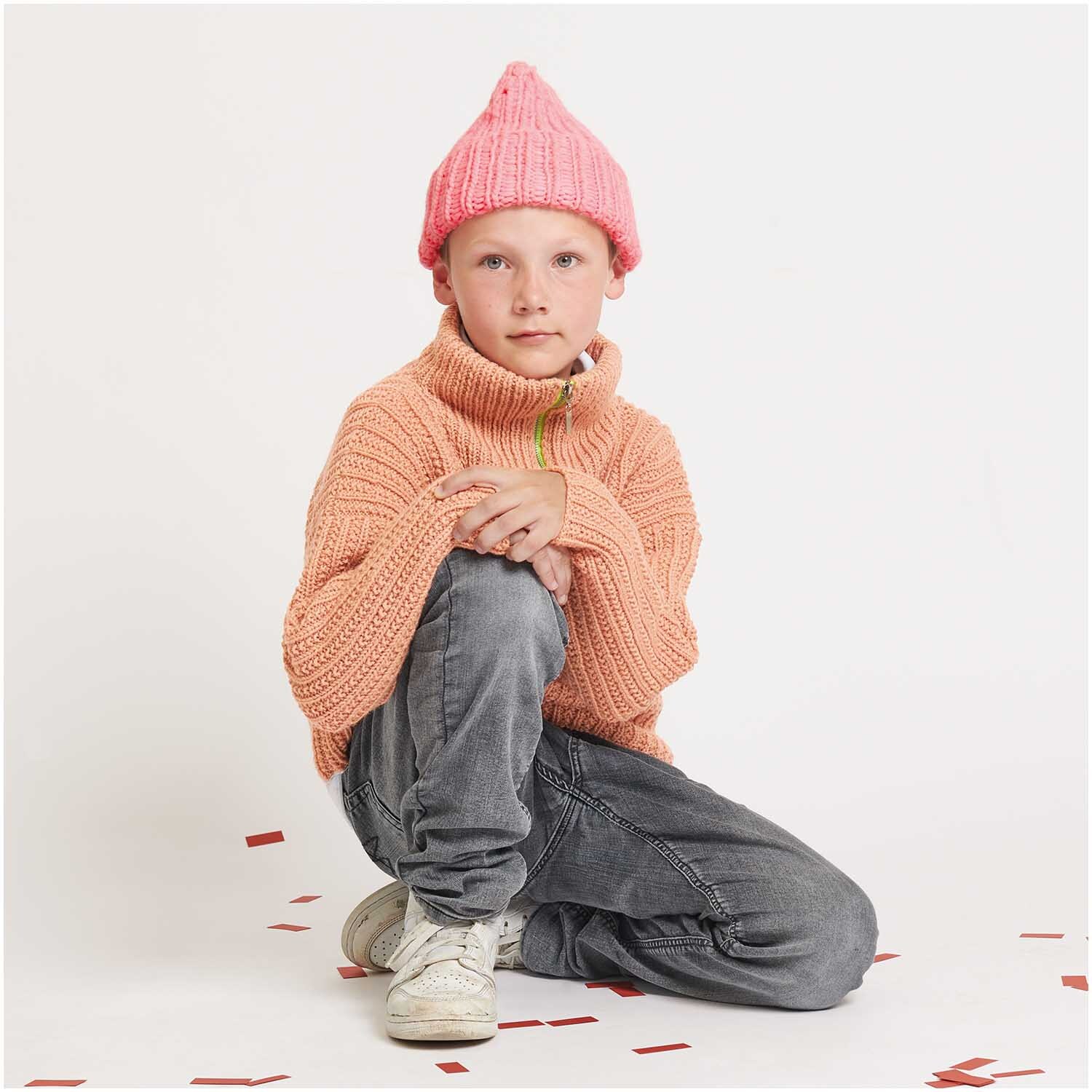 Strickset Mütze Modell 21 aus Kids Nr. 12: Kind mit rosa Mütze und orangefarbenem Strickpullover.