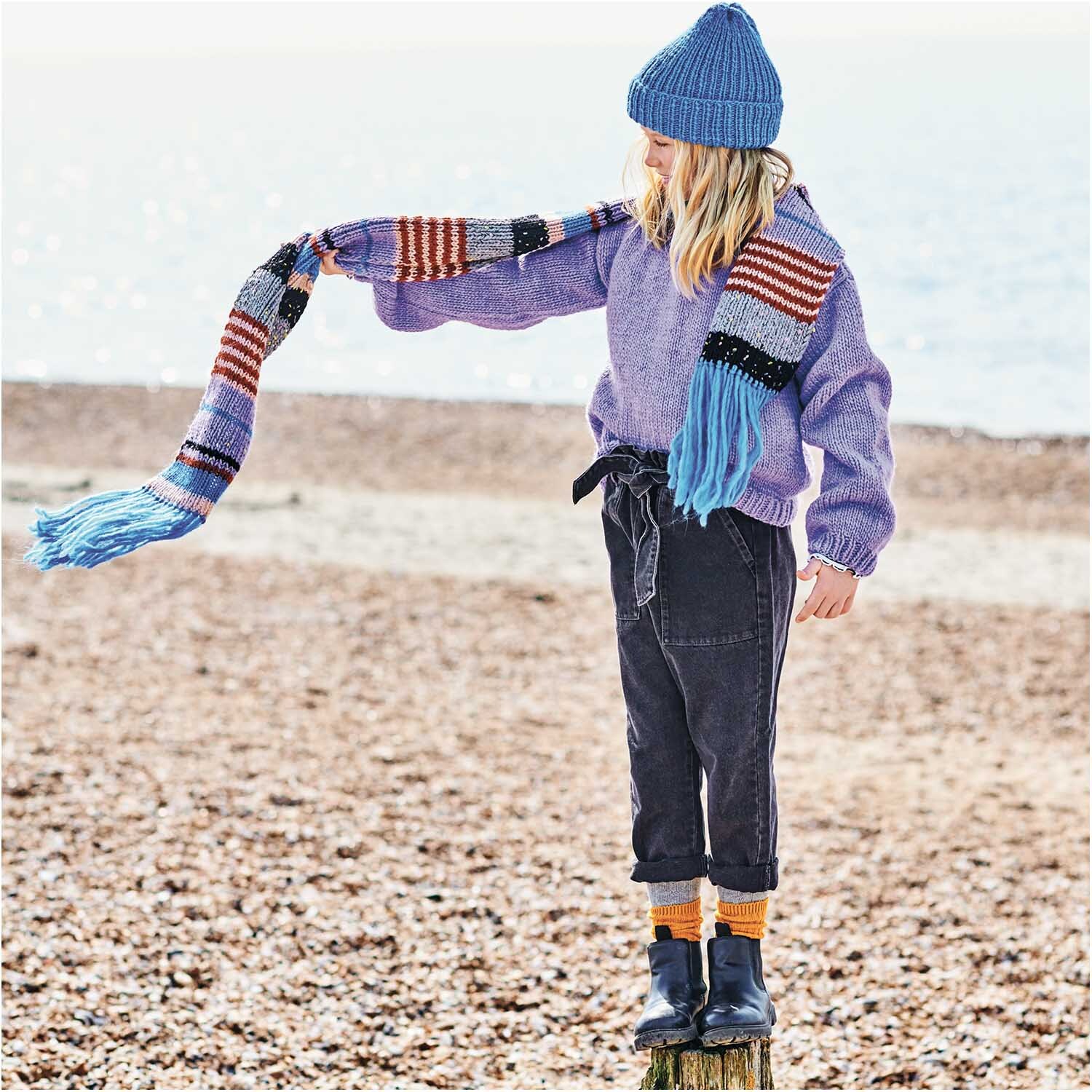 Essentials Super aran 50g 100m Farbe Rost von Rico Design Essentials Super aran: Kind in lila Pullover und bunter Schal am Strand, trägt blaue Mütze und schwarze Stiefel.