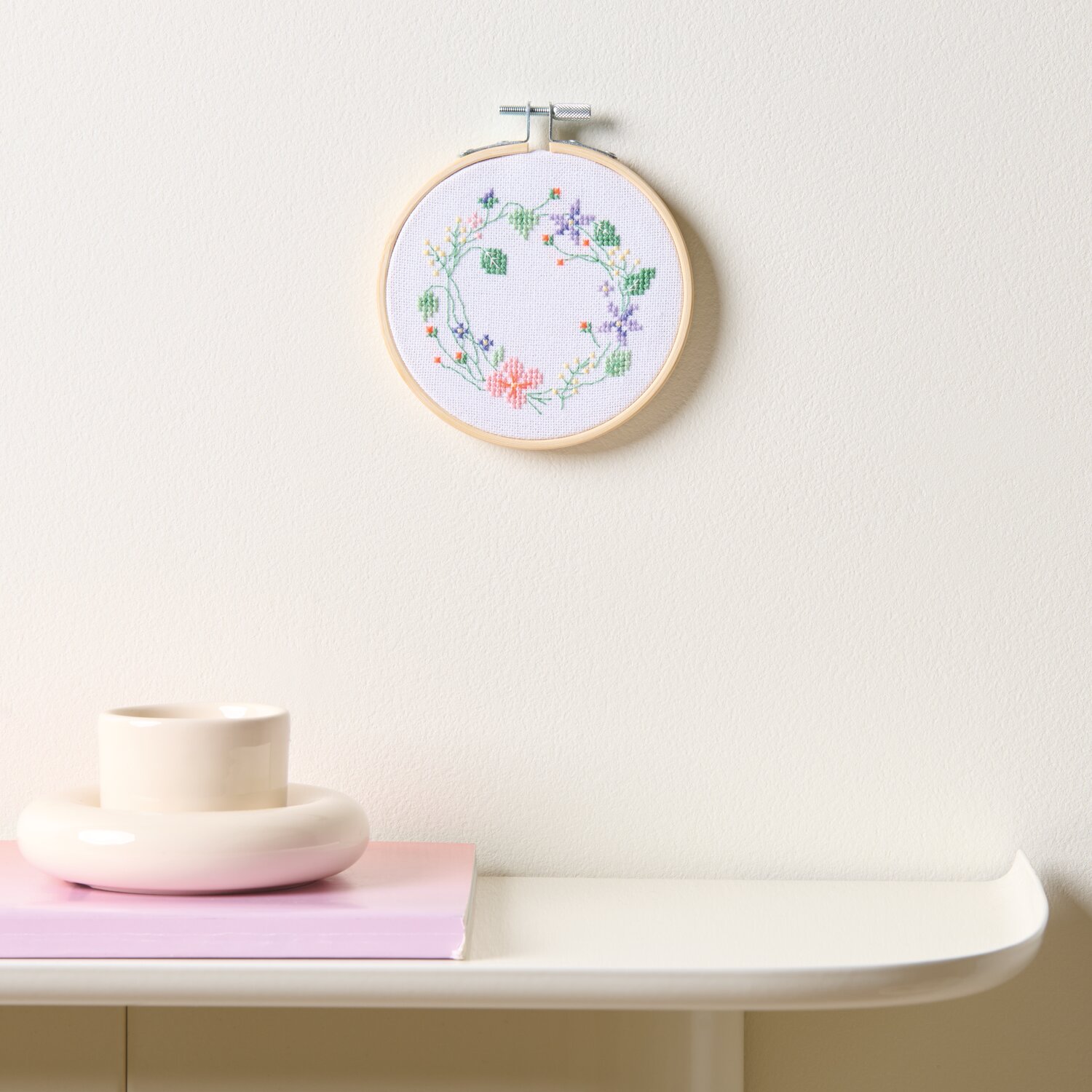 Stickpackung Gezählter Kreuzstich Blumenkranz, Ø10cm, an weißer Wand über Tisch mit Deko.