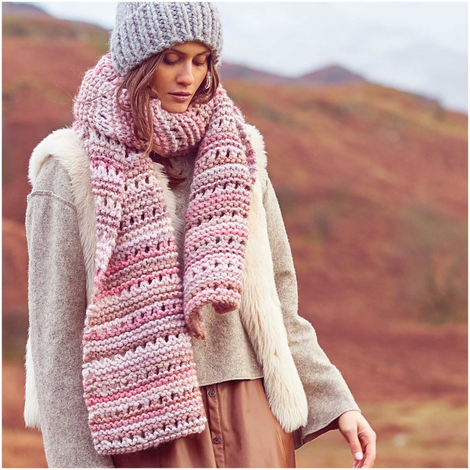 Creative Smile: Frau trägt gestrickten Schal und Mütze in Rosa von Rico Design, umgeben von herbstlicher Landschaft.