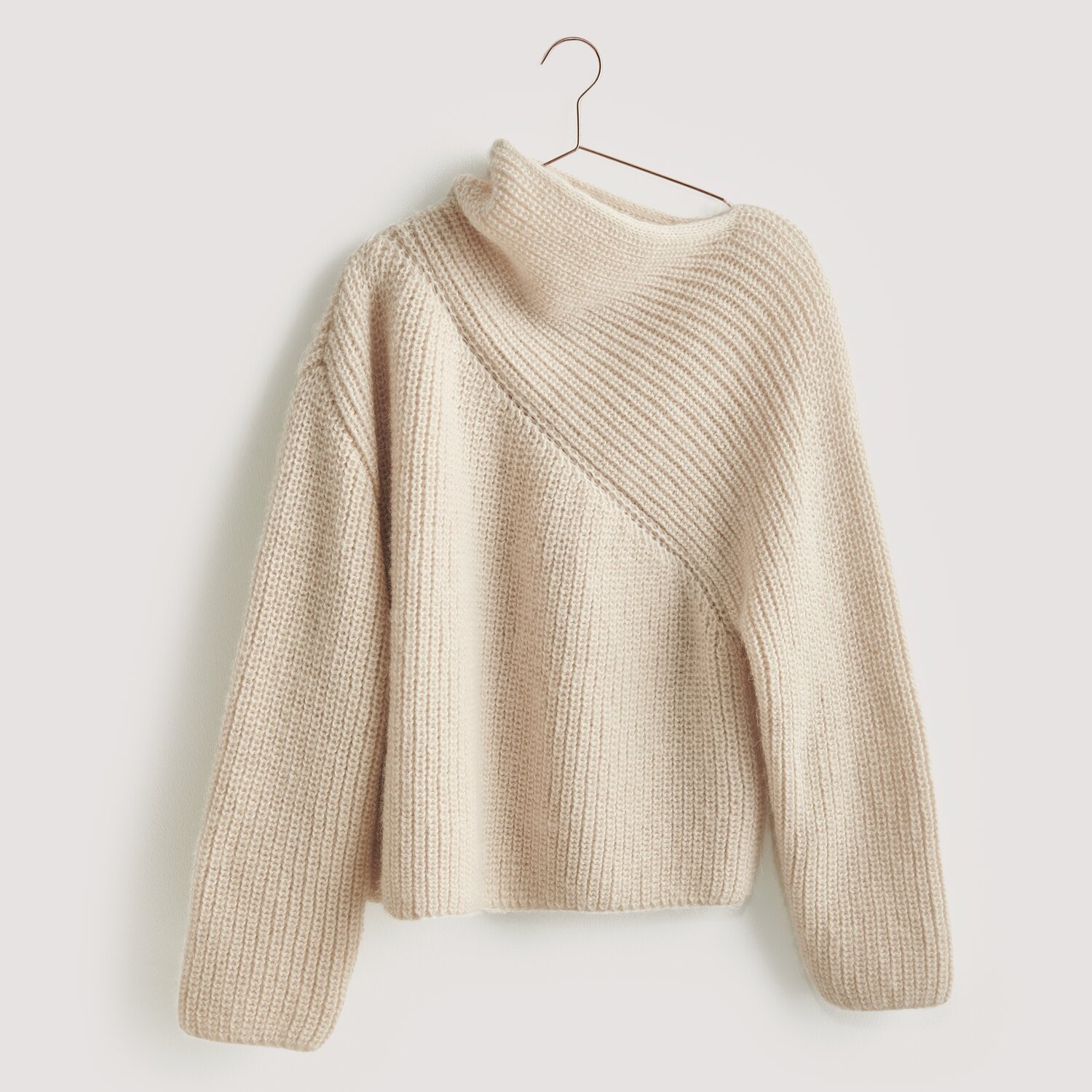 Strickset Pullover Modell 01 aus Luxury Knits: Beigefarbener Pullover mit asymmetrischem Strickmuster, hängend an einem Kleiderbügel.