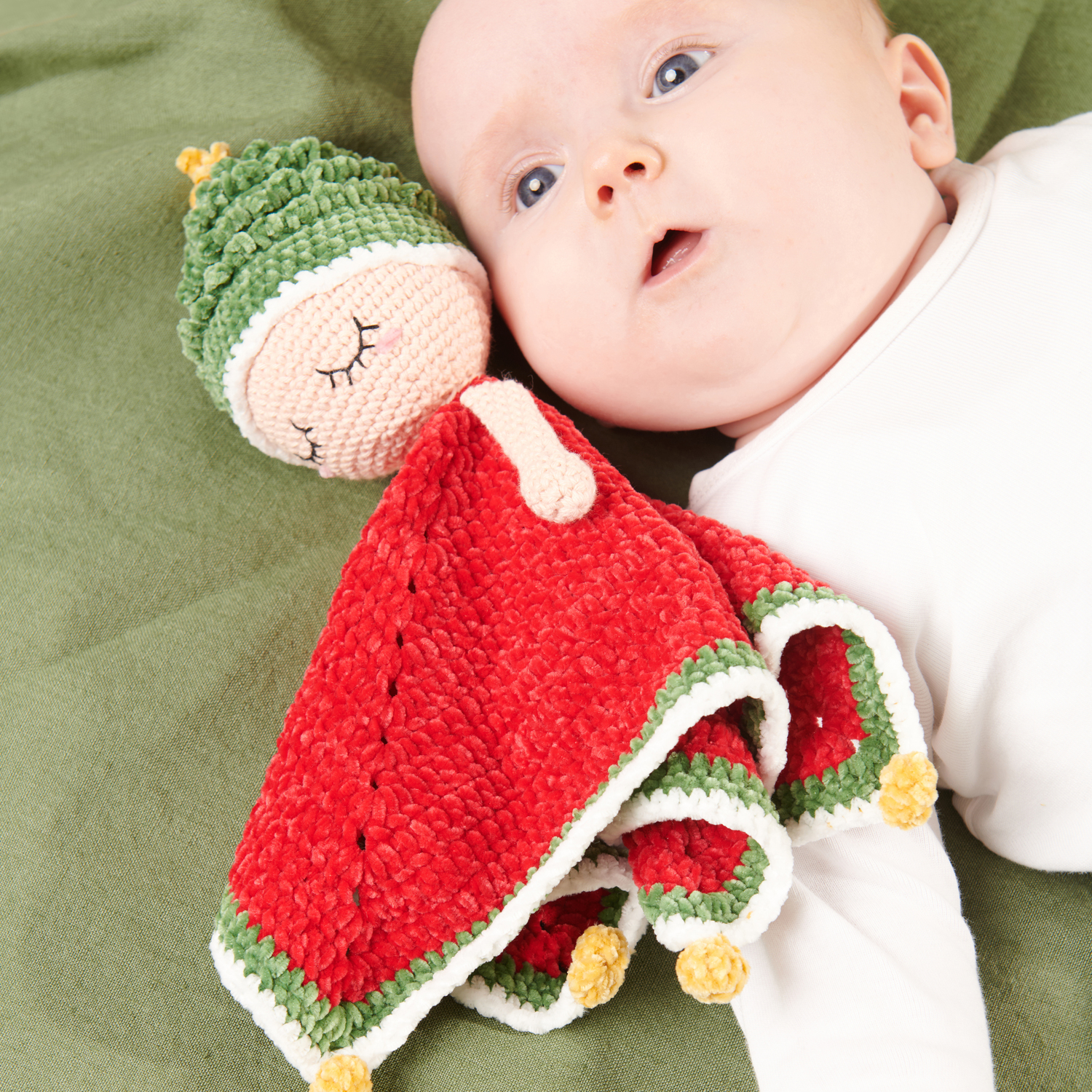Häkelset Ricorumi Baby Blankies Weihnachtswichtel: Baby mit rotem und grünem Häkelwichtel auf grünem Hintergrund.