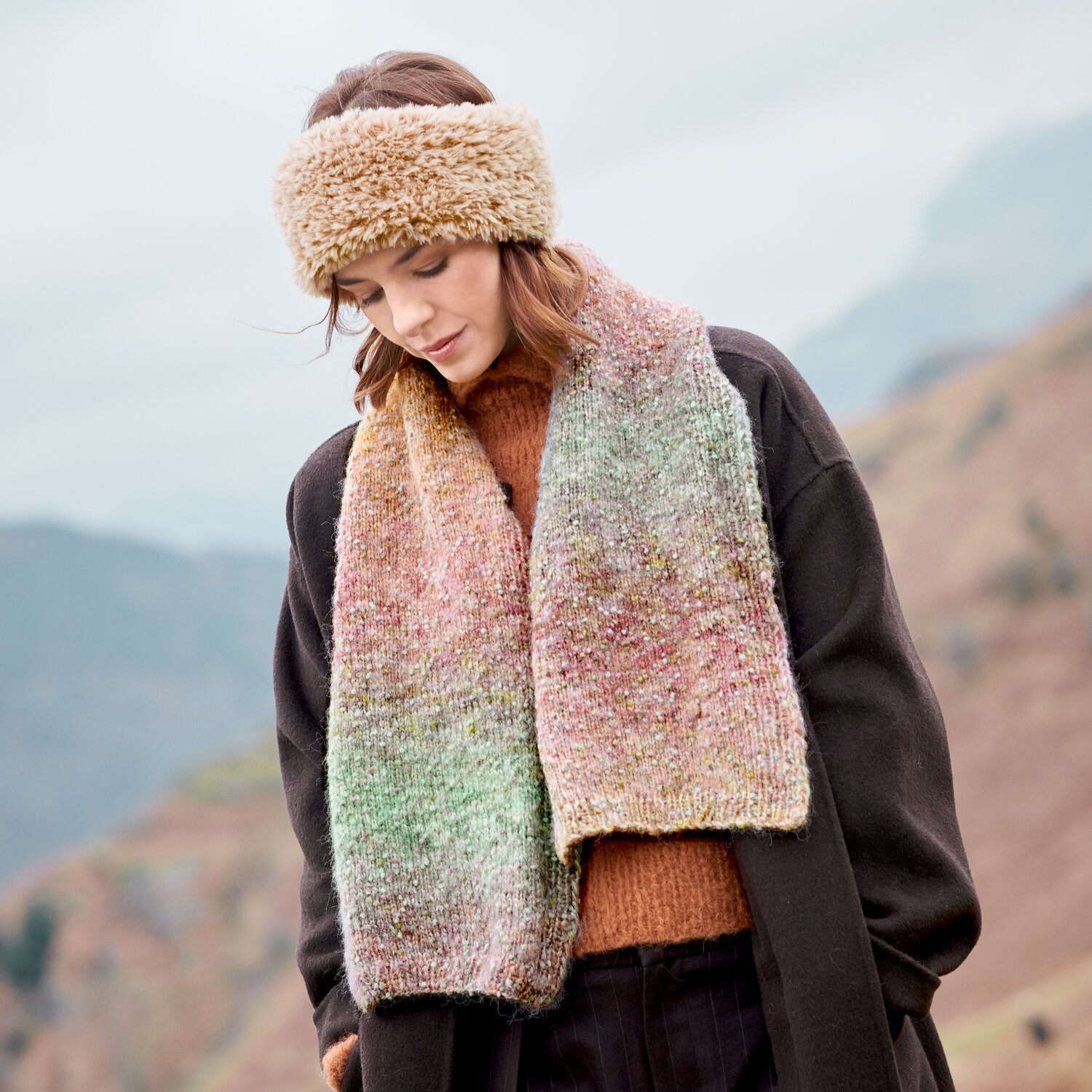 Strickset Stirnband Modell 26 aus Lovewool No. 23, getragen von Frau mit flauschigem Stirnband und langem, mehrfarbig meliertem Schal in herbstlicher Berglandschaft