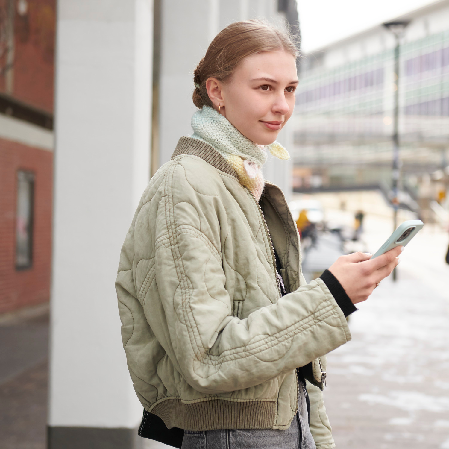 Strickset Mini Dreieckstuch Modell 01a: Frau mit hellgrüner Jacke und Smartphone in urbaner Umgebung.