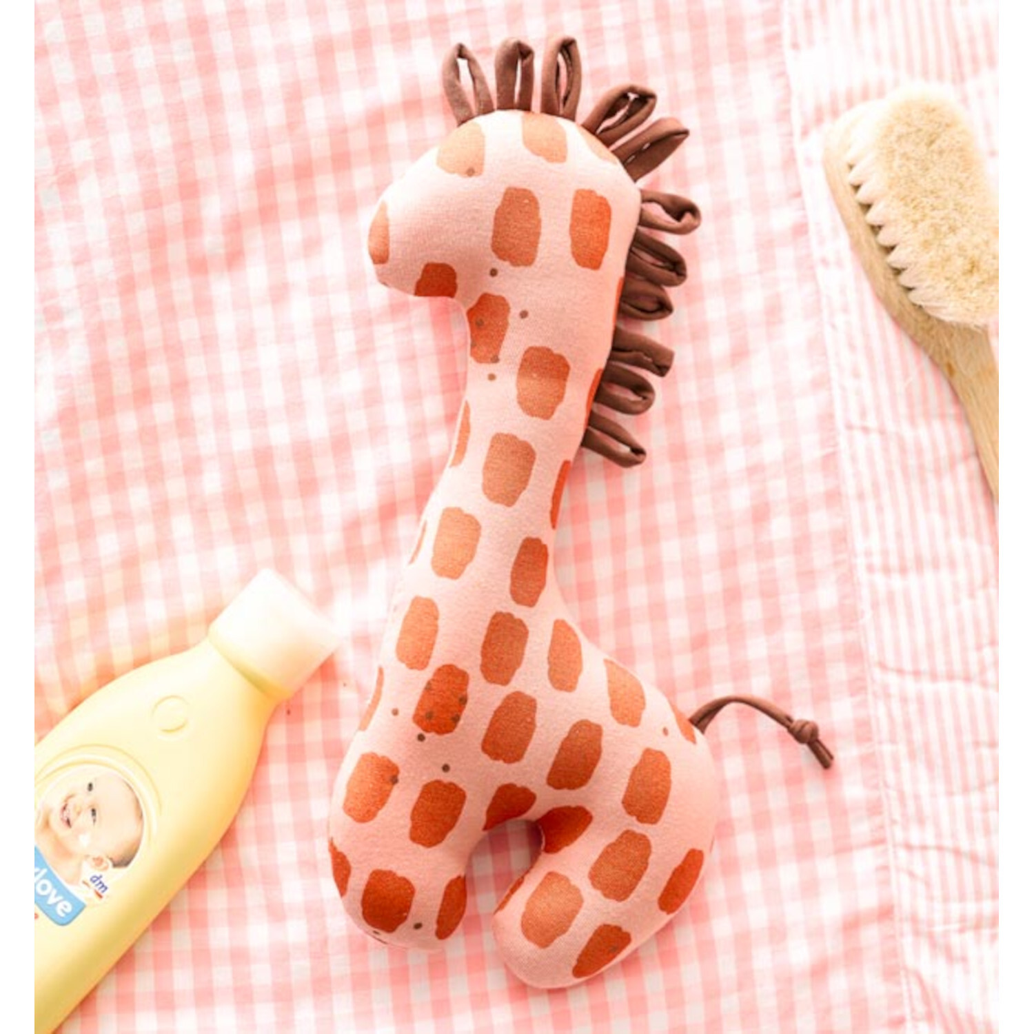 Nähen mit Jersey für Babys: Rosa Stoffgiraffe auf kariertem Hintergrund, neben Babyshampoo und Bürste.