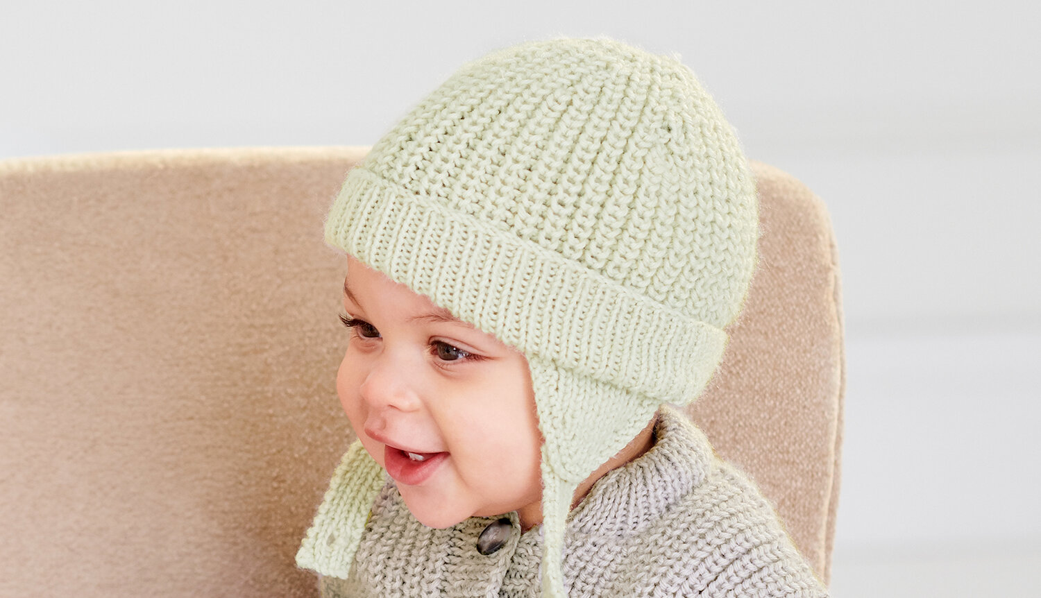 Strickanleitung Mütze und Strampler aus Rico Baby Dream dk uni, lachendes Baby mit weicher Strickmütze mit Ohrenklappen und passendem Oberteil auf hellem Sessel
