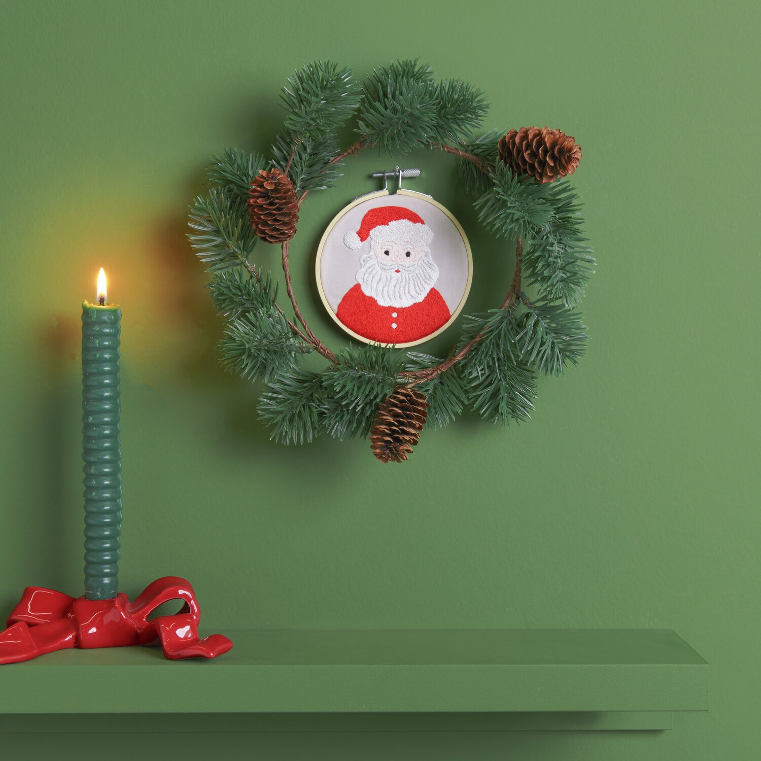Stickpackung Santa vorgezeichnet: Ein weihnachtlicher Kranz mit Tannenzweigen und einem gestickten Santa-Motiv, dekoriert mit Zapfen und einer grünen Kerze.