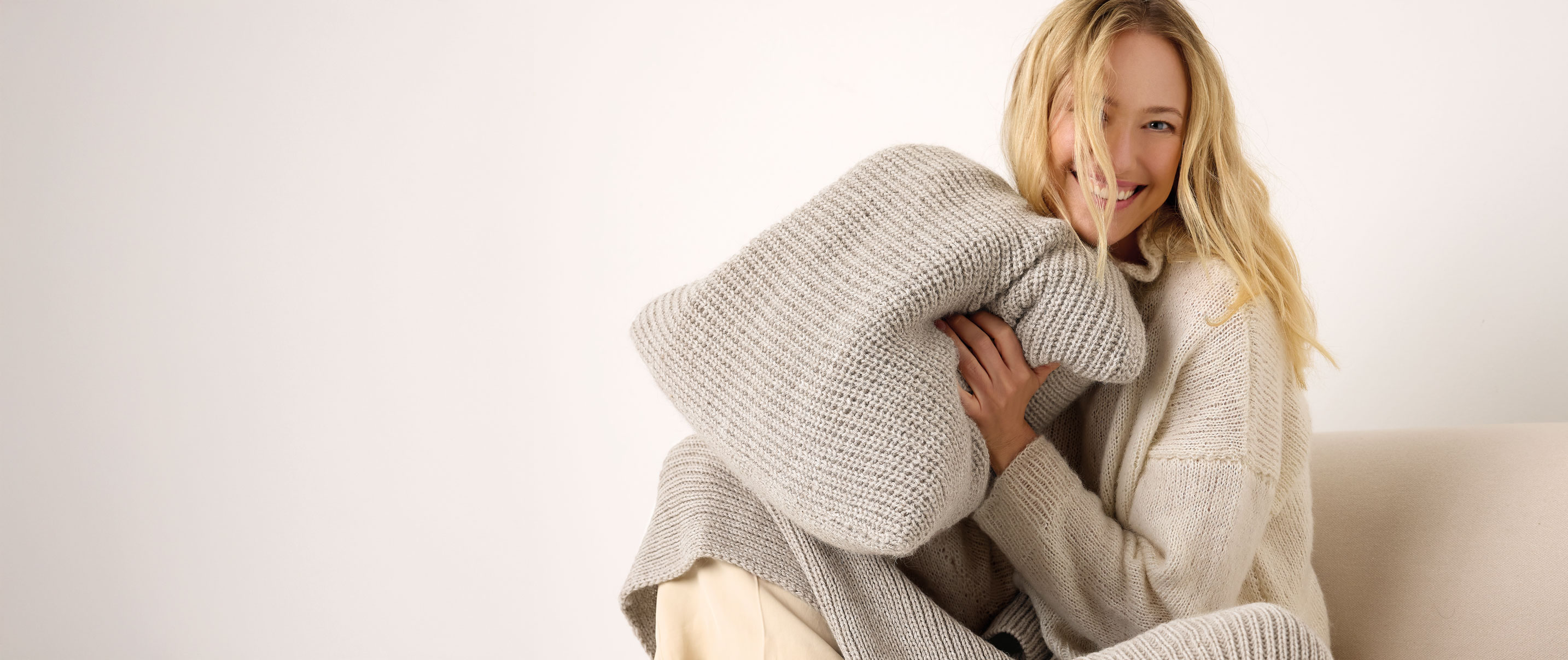 Frau hat graues Strickkissen in der Hand und sitzt mit Strickdecke auf einem Sofa