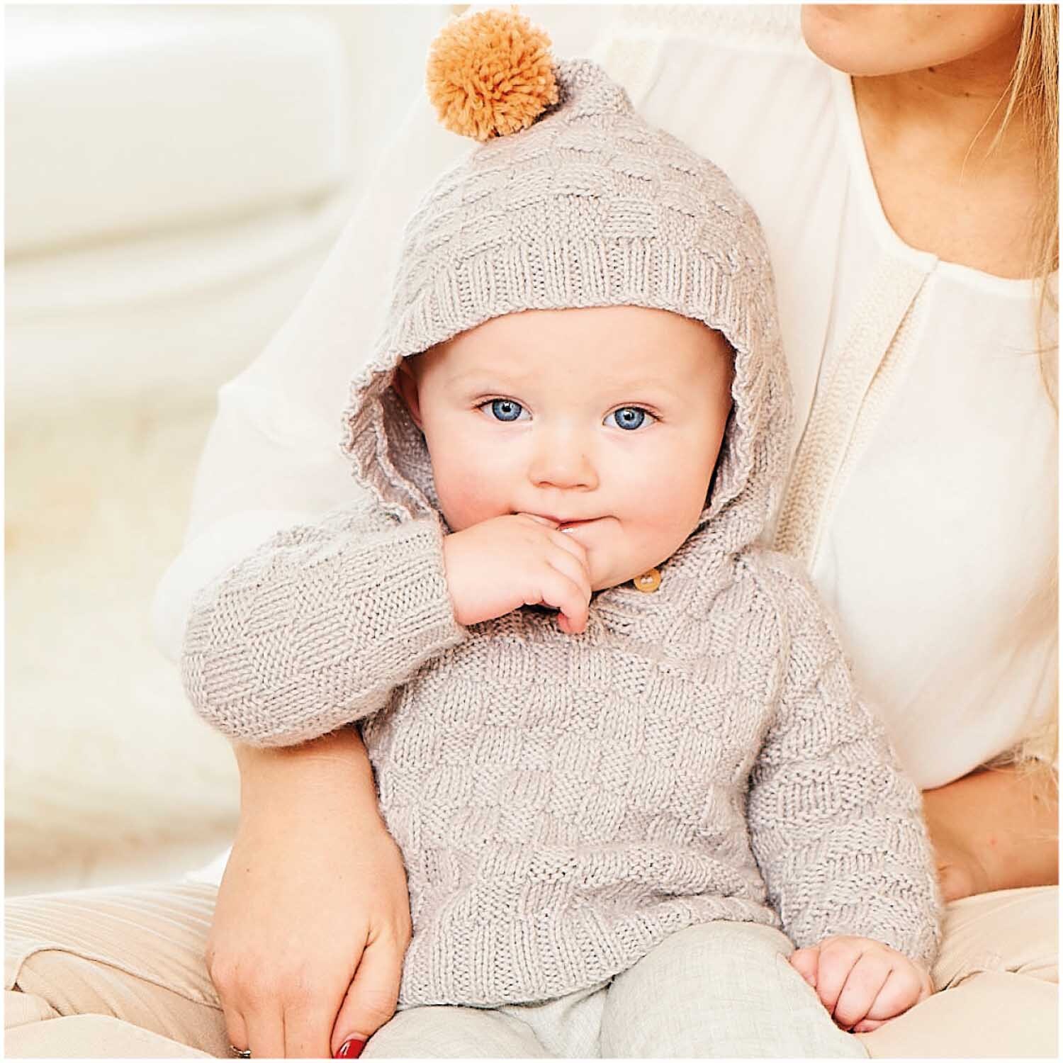 Rico Baby Dream dk uni - Ein Baby in einem gestrickten Hoodie in Silbergrau mit Kapuze und Bommel, gehalten von einer Frau.